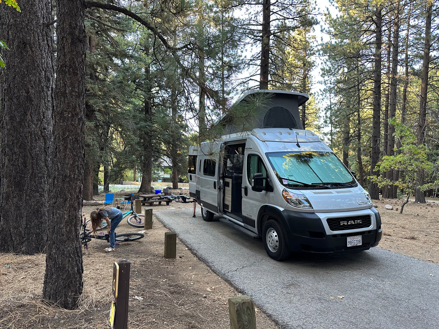 RV rental parked at a shaded forest campsite. A person works on a bike near the pop-top camper van, ready for outdoor adventures.