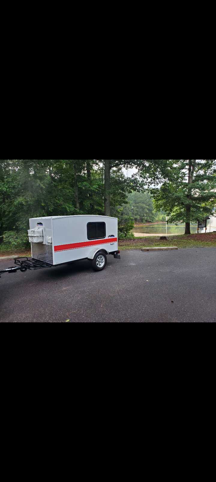 Small white travel trailer parked by a lake, surrounded by green trees. Perfect for your next rv rental adventure.