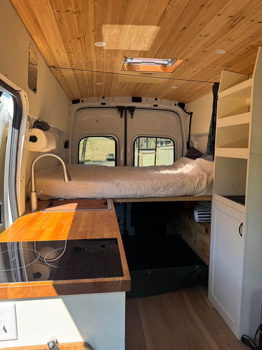 Cozy rv rental interior: wood ceiling, bed, kitchenette w/ induction cooktop & sink. Perfect for van life adventures.