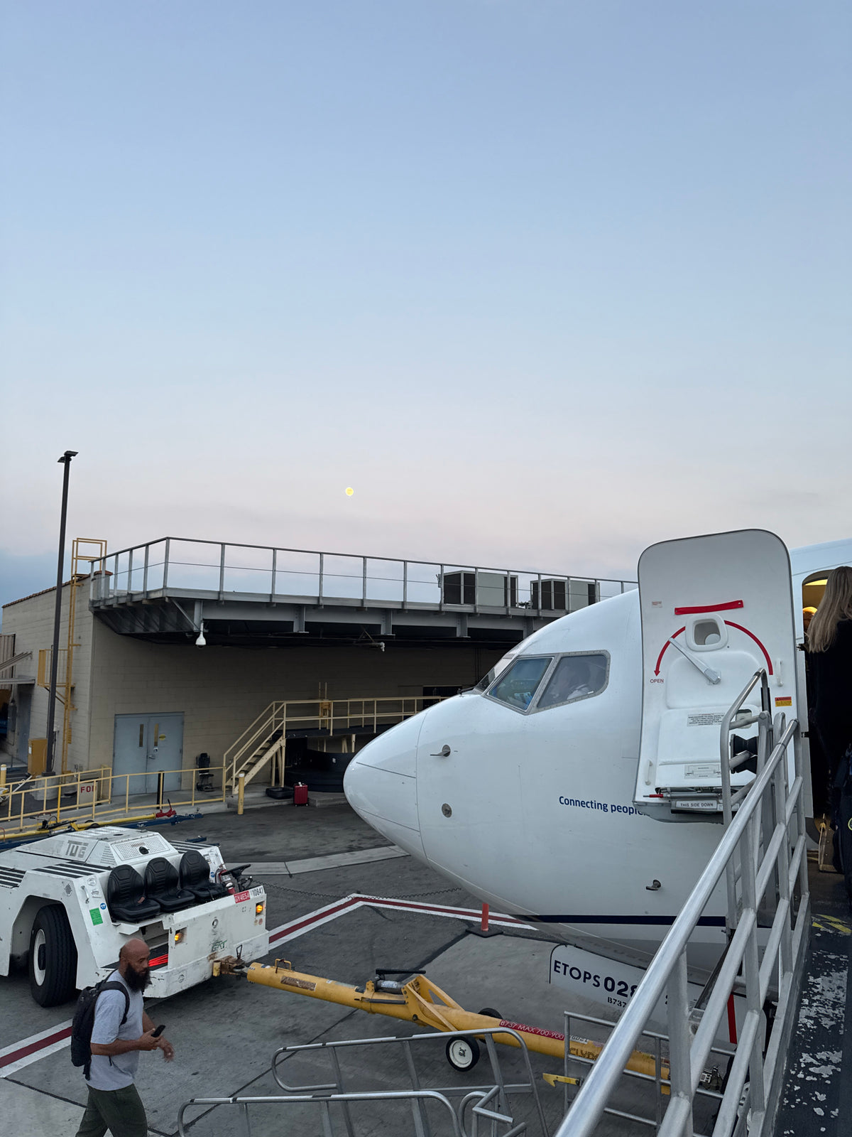 A large white airplane at an airport gate, with people boarding via stairs. A tow tug is nearby. Unlike an **rv rental**, this is air travel. Moon visible overhead. — photo 11