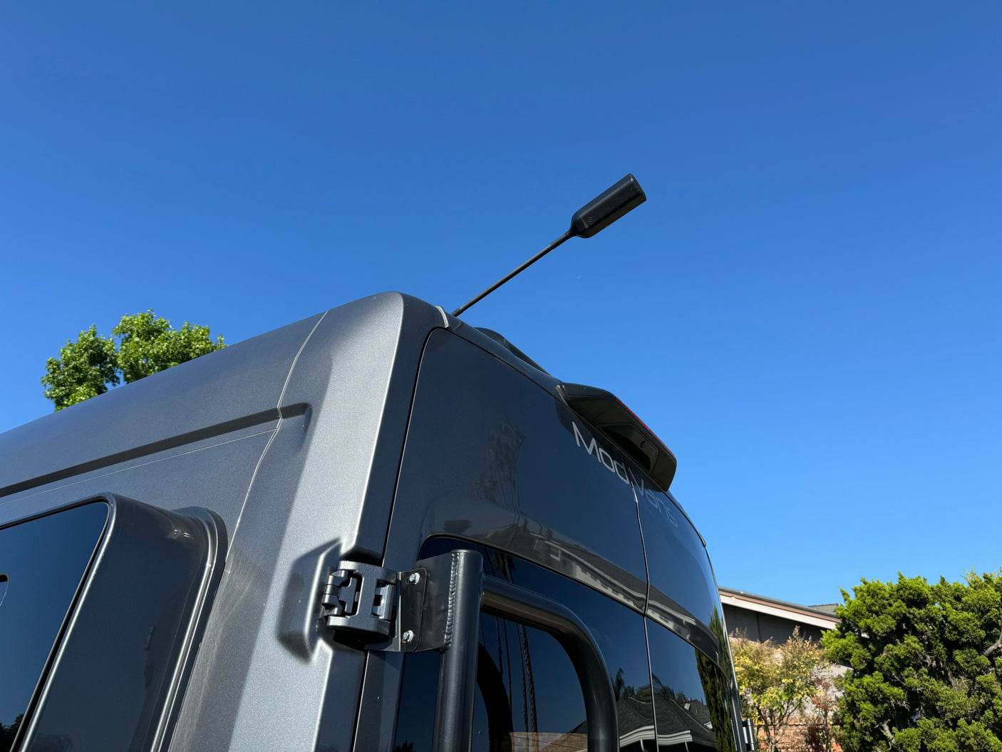 Up-close rear view of a sleek gray ModVans camper van, showing its roof antenna, spoiler, and a ladder, available for RV rental.
