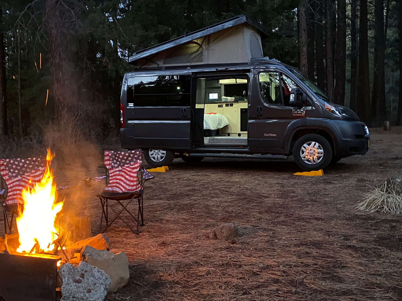 Enjoying a cozy evening camping with an RV rental. A dark grey pop-top camper van, American flag chairs, and a bright campfire at dusk.