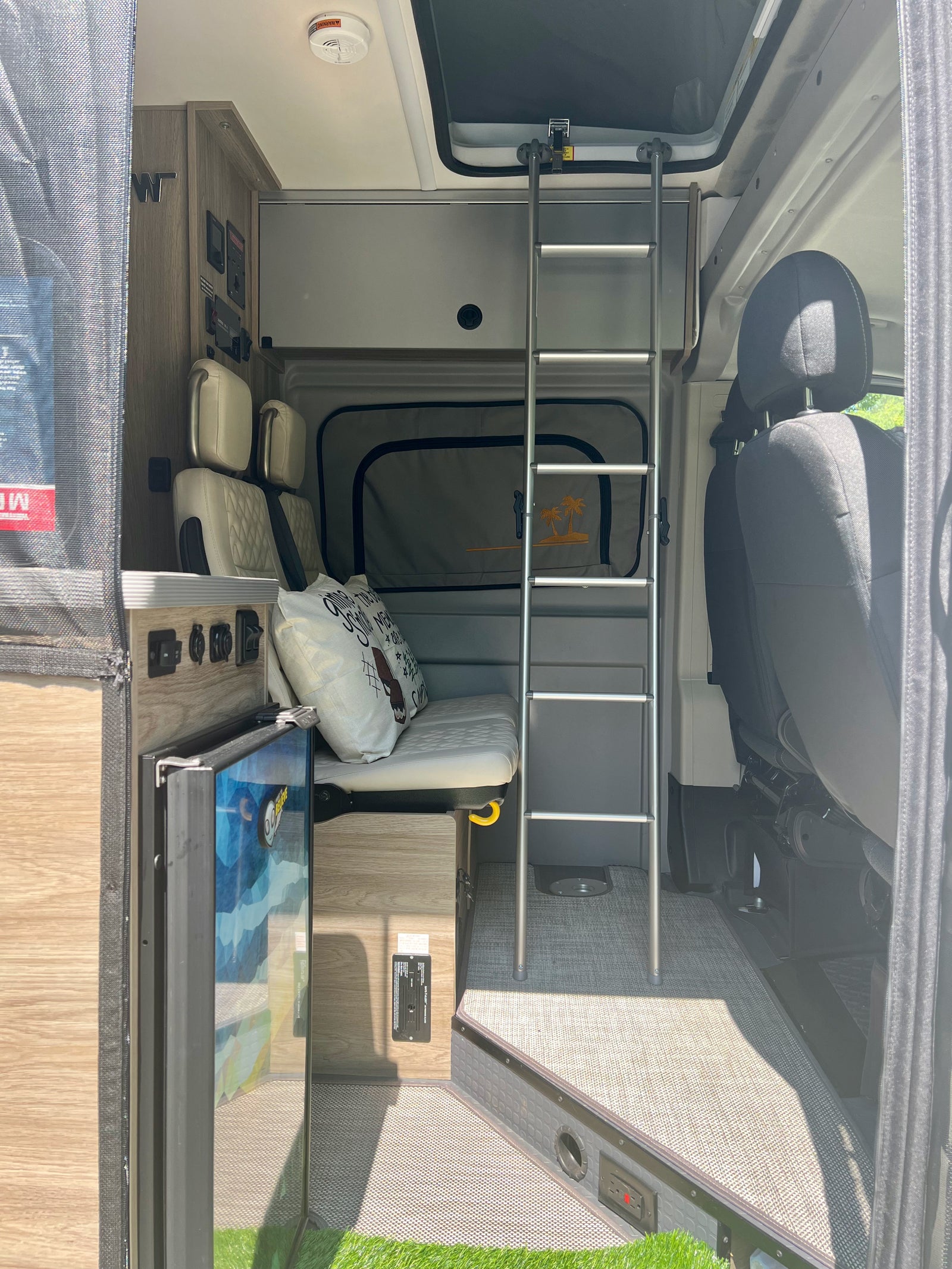 View inside a modern RV rental featuring a bunk bed, bench seating, compact fridge, and control panel for road trip comfort.