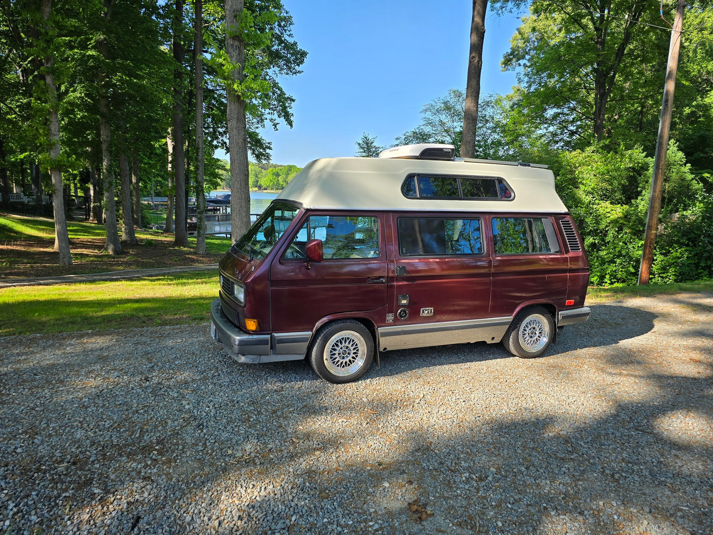 Maroon VW Vanagon camper van on gravel by a lake and trees