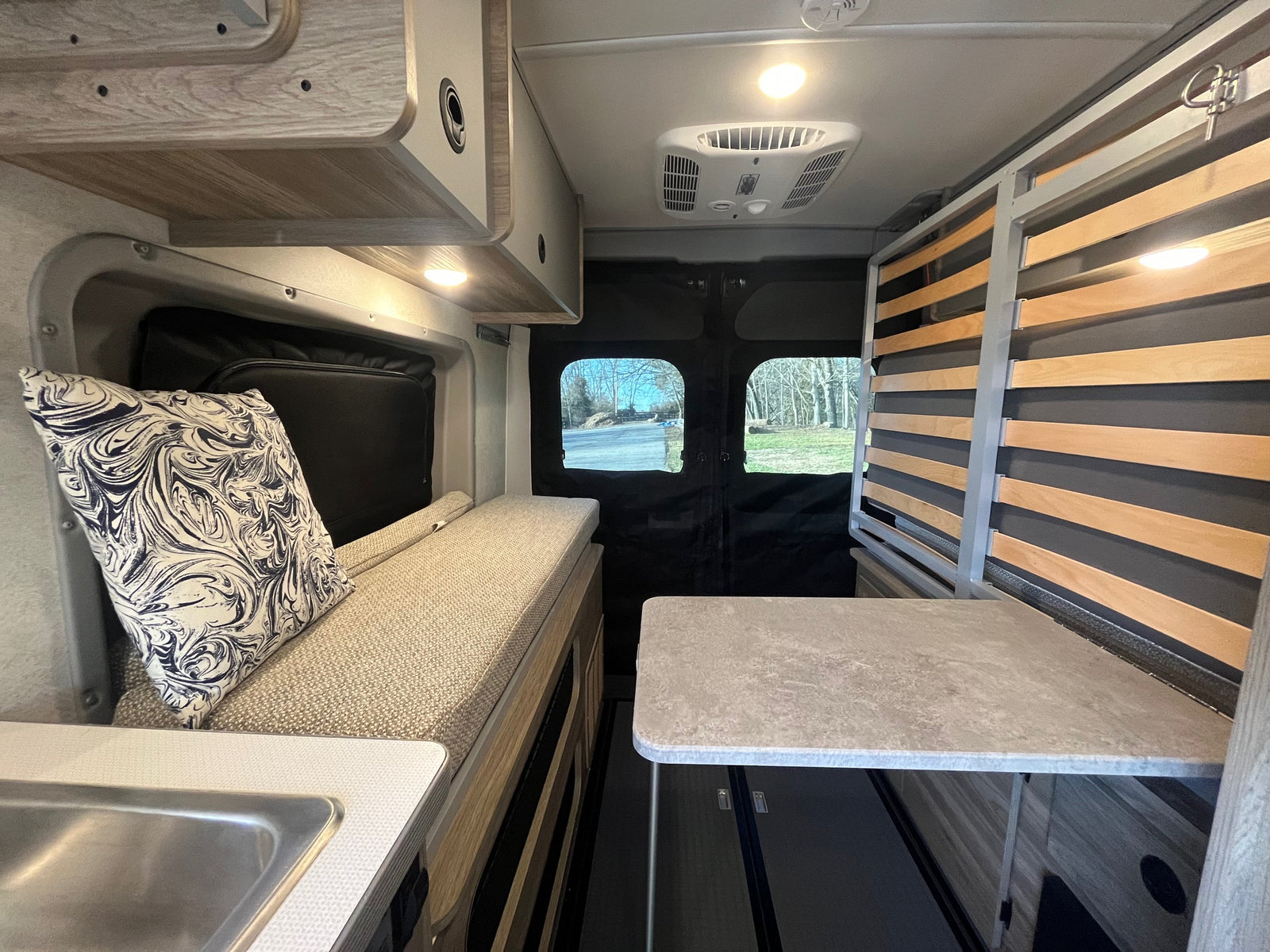 This rv rental interior has a cozy bench, sink, table, and slatted bed frame, with bright overhead lights and outdoor views.