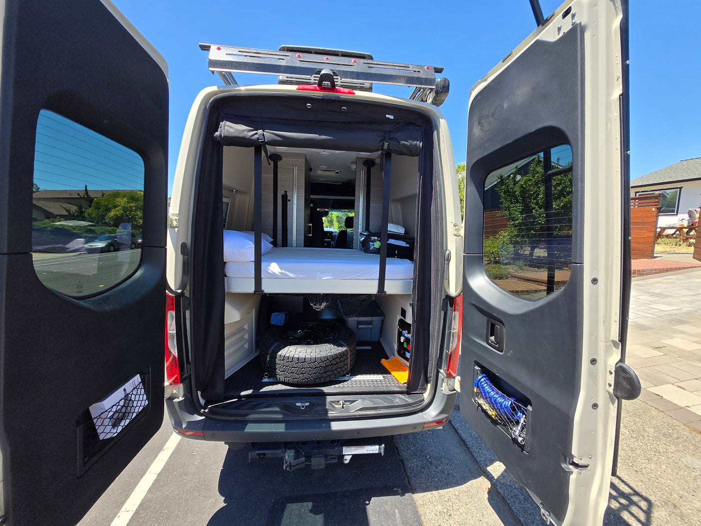 RV rental: Open rear doors reveal a versatile camper van interior with a bed, under-bed storage, and utility access.