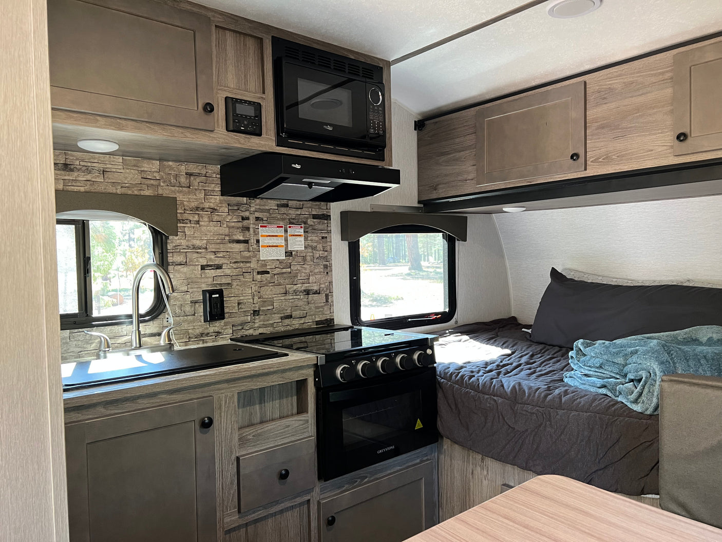 Explore this compact RV rental, showcasing a modern kitchen with a microwave, stove, sink, and a cozy bed area.