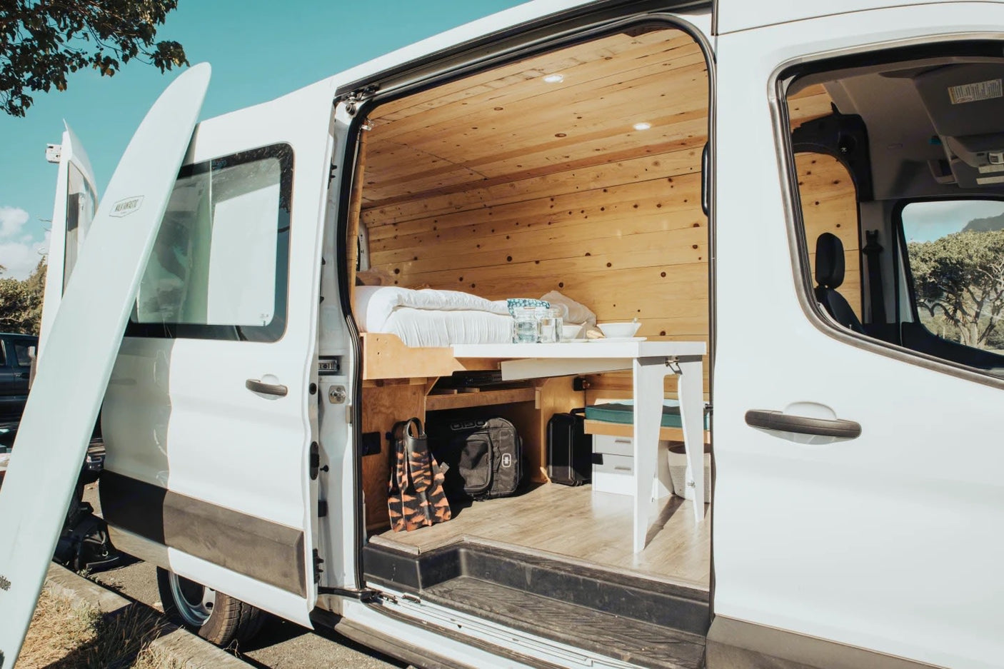 This RV rental shows a white camper van with its side door open, revealing a cozy wooden interior with a bed, table, and a surfboard outside.