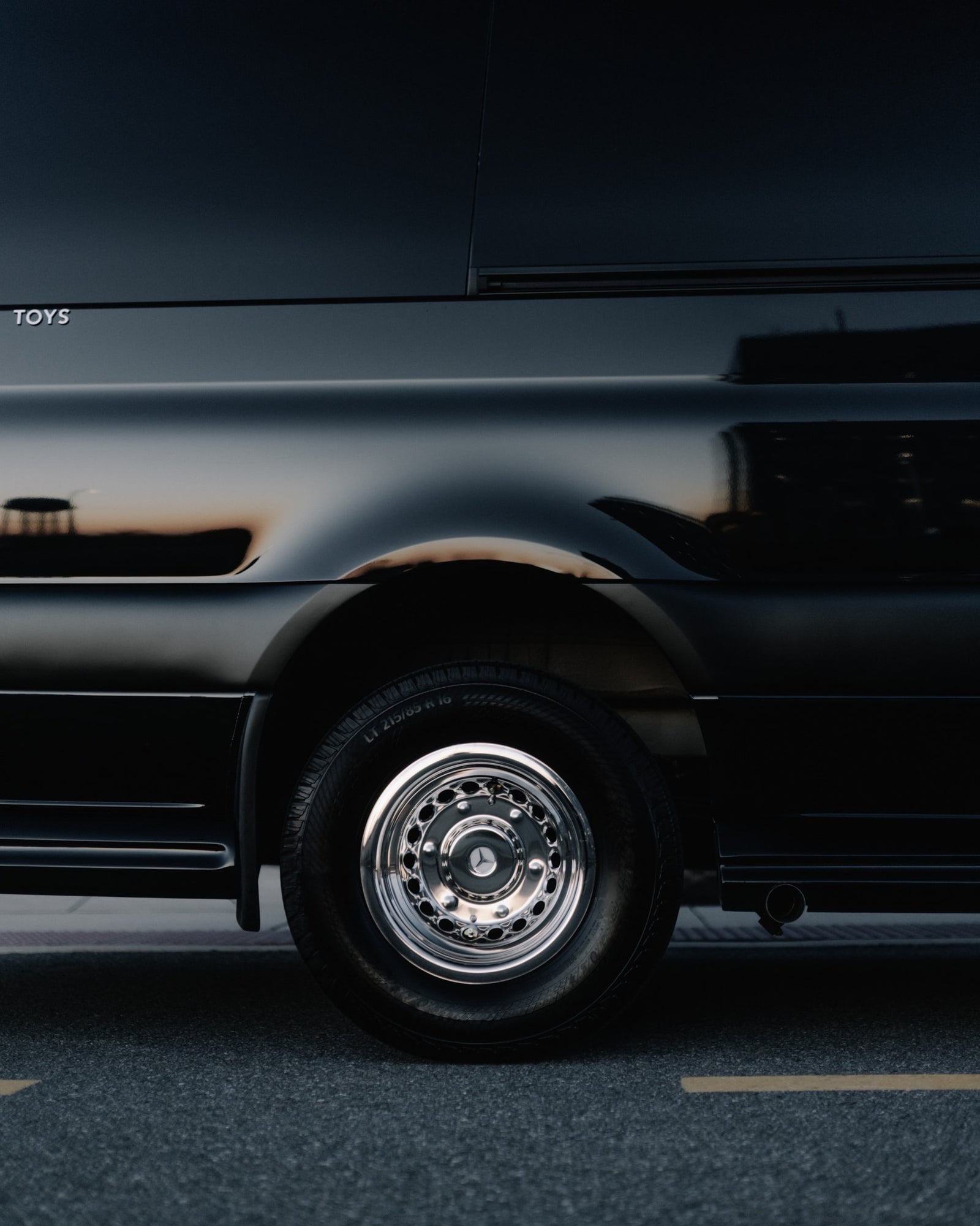 Black luxury RV rental side view with chrome wheel, Mercedes logo, and warm reflections. "TOYS" visible.