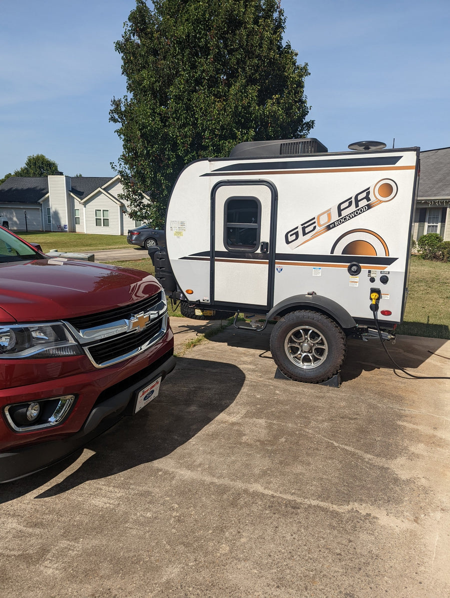 Red Chevy truck hitched to a white Geo Pro travel trailer, likely an RV rental, parked on a driveway on a sunny day.