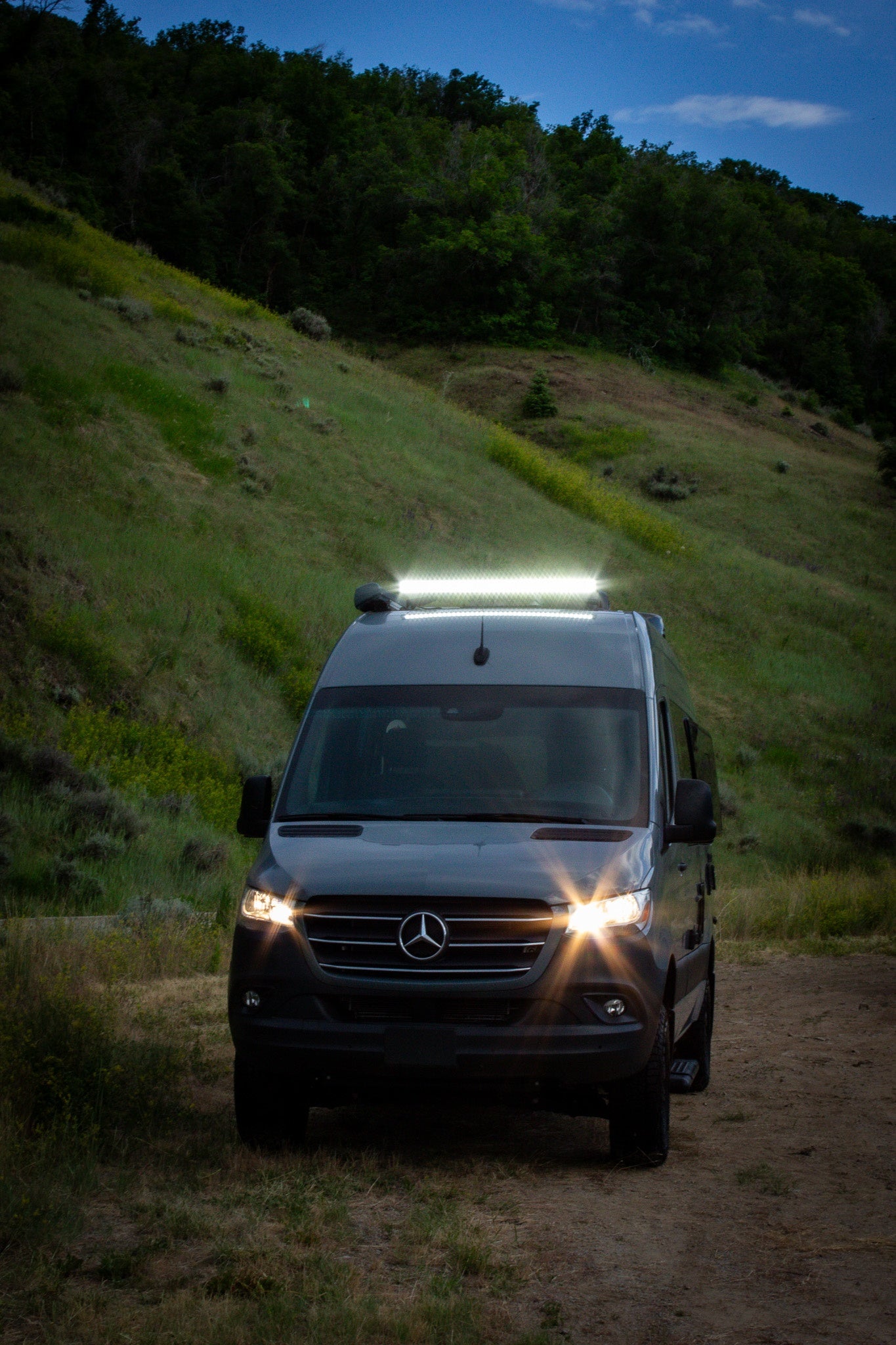 This dark grey Mercedes Sprinter camper van, a bright RV rental, illuminates a dirt path at dusk in a hilly landscape.