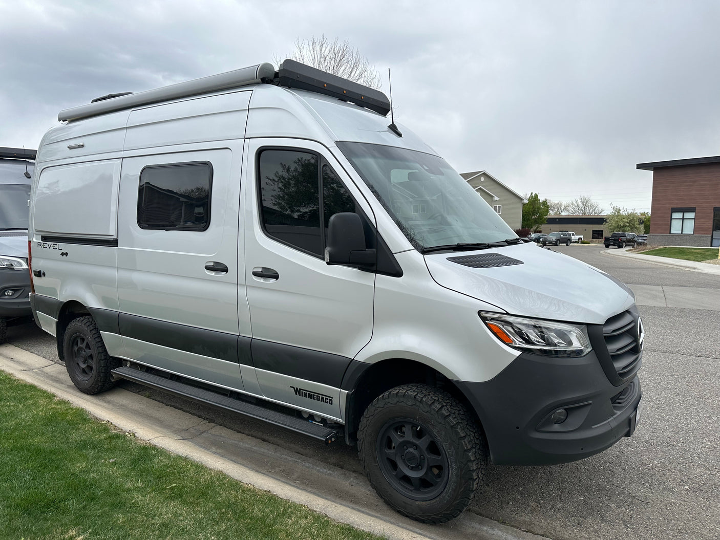 A silver Winnebago Revel camper van, an ideal rv rental, with rugged tires and roof awning, parked on a street.