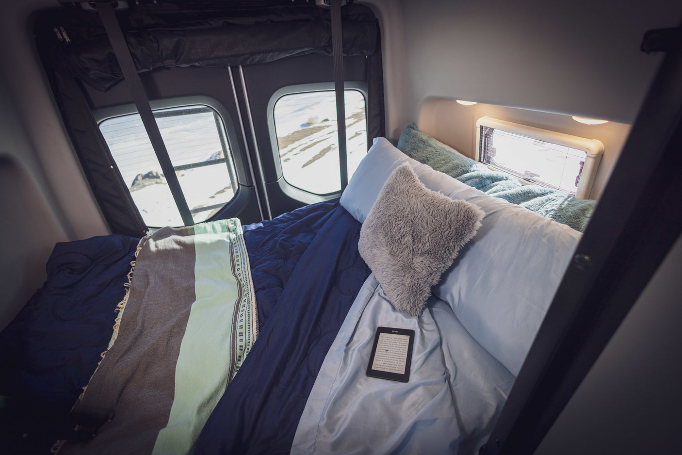 Cozy RV rental bed with a view of a snowy landscape and an e-reader, perfect for a winter escape in a camper van.