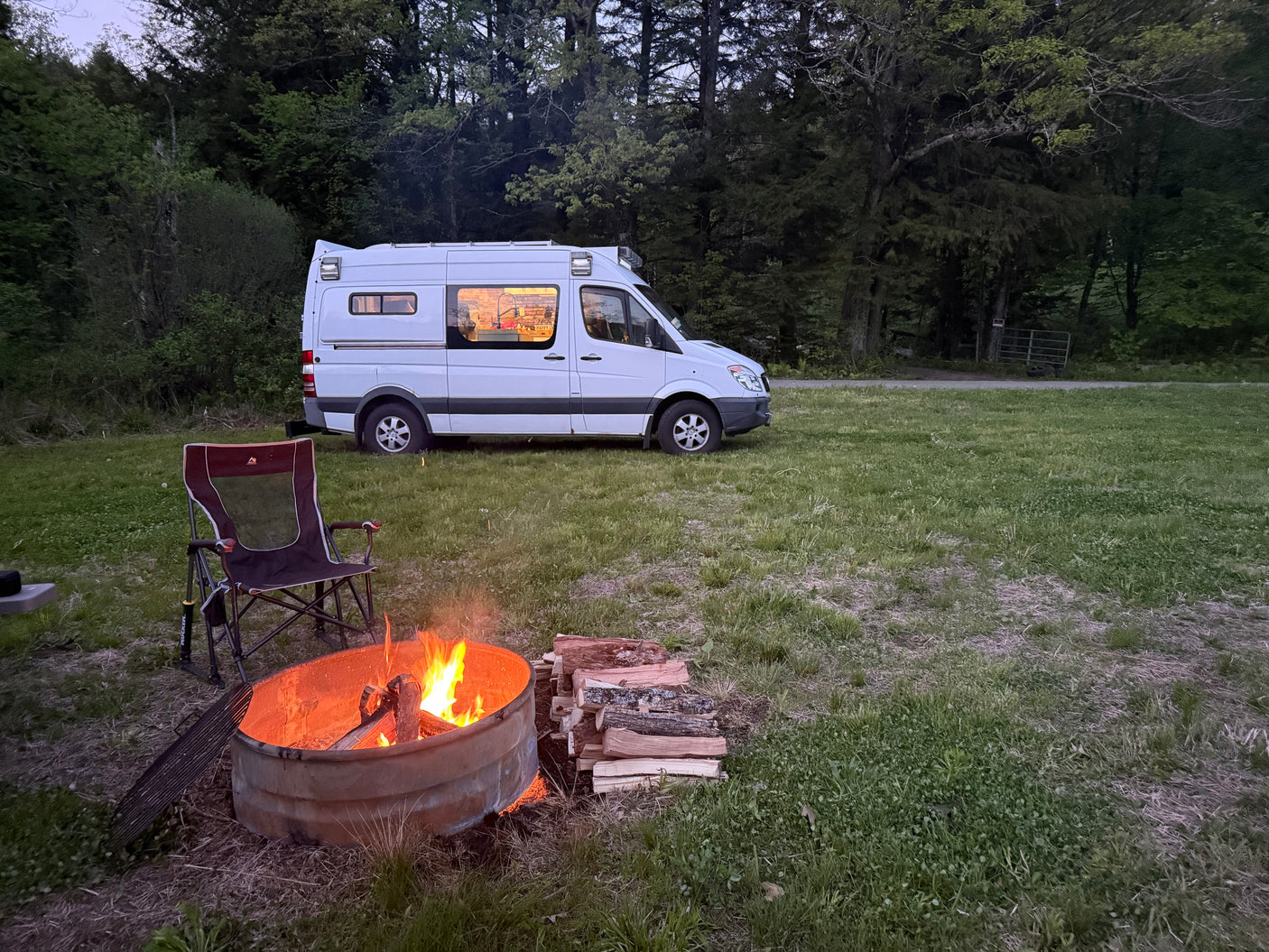 Cozy night camping with an RV rental: a white camper van, crackling campfire, and a chair on green grass near trees.