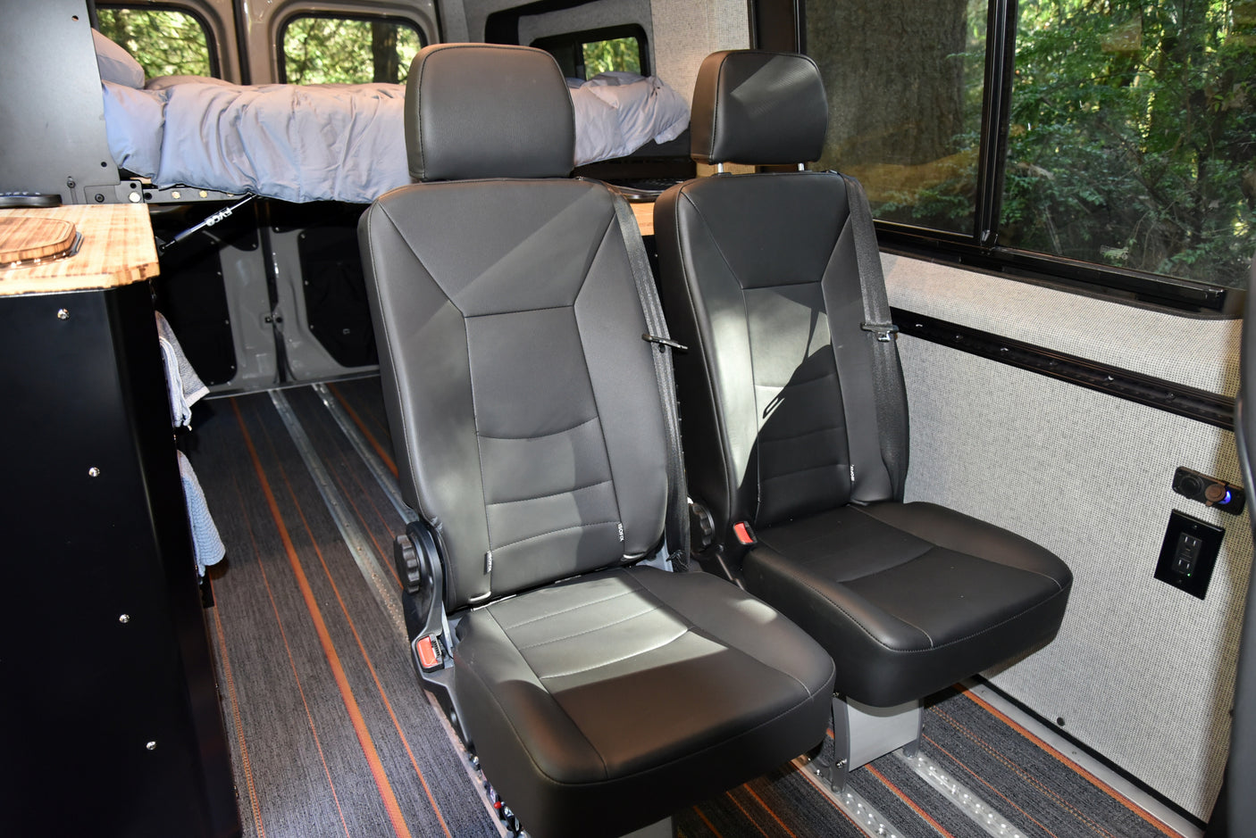 Inside this RV rental: two black passenger seats and an elevated bed, providing flexible seating & sleeping in a camper van.