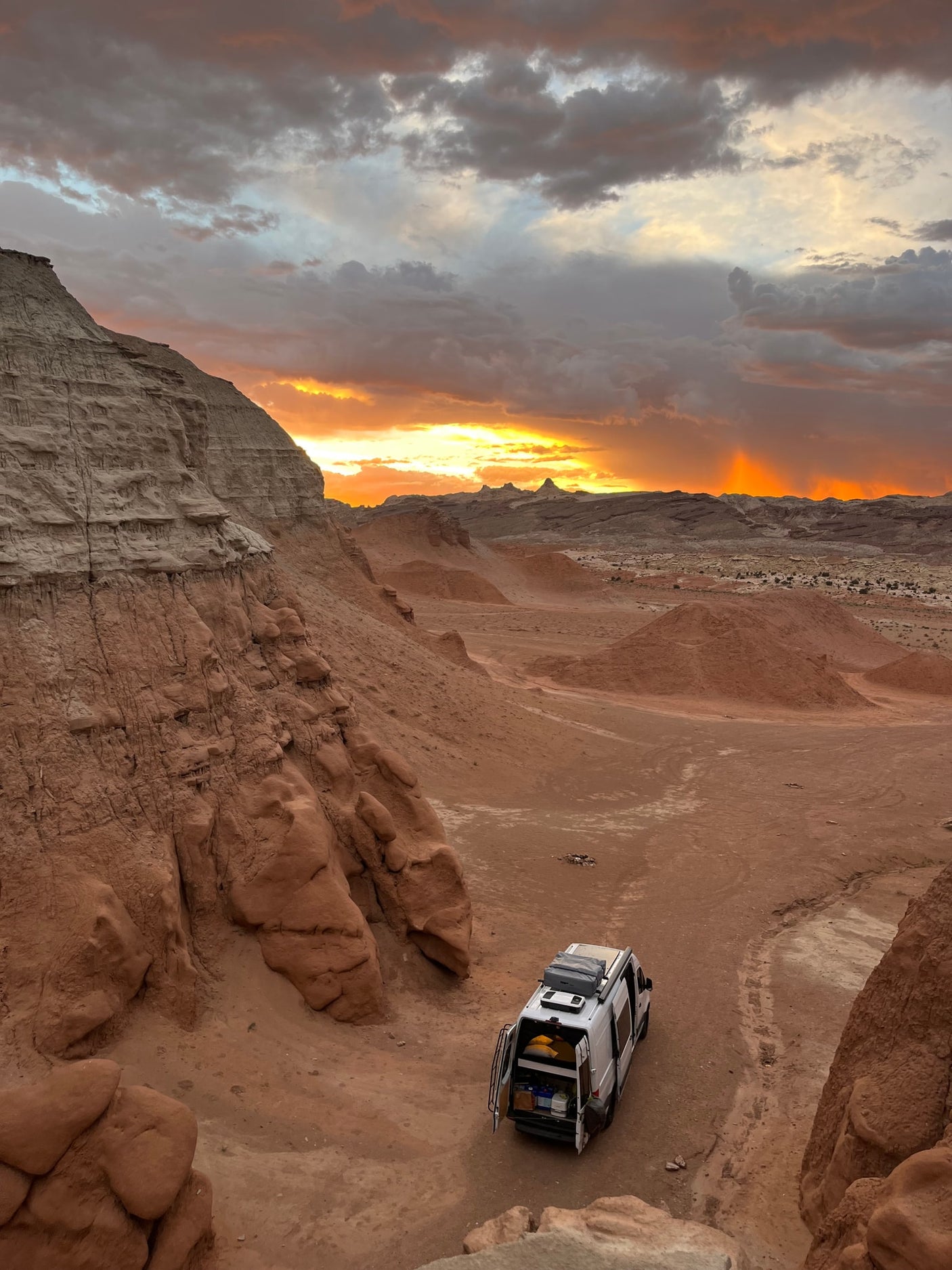 Golden sunset lights a white RV rental parked in a rugged desert valley with towering cliffs. A perfect spot for remote adventure.