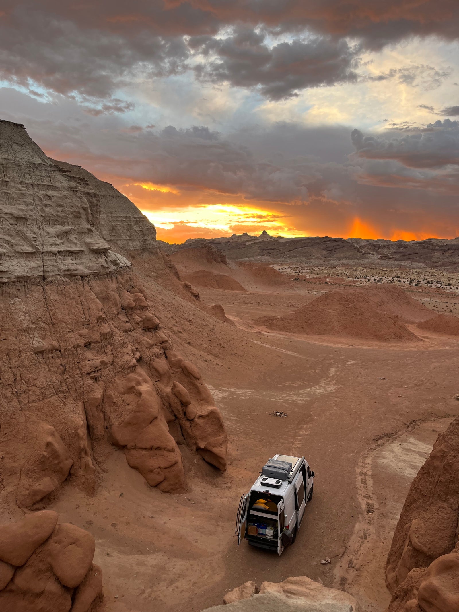 Golden sunset lights a white RV rental parked in a rugged desert valley with towering cliffs. A perfect spot for remote adventure.