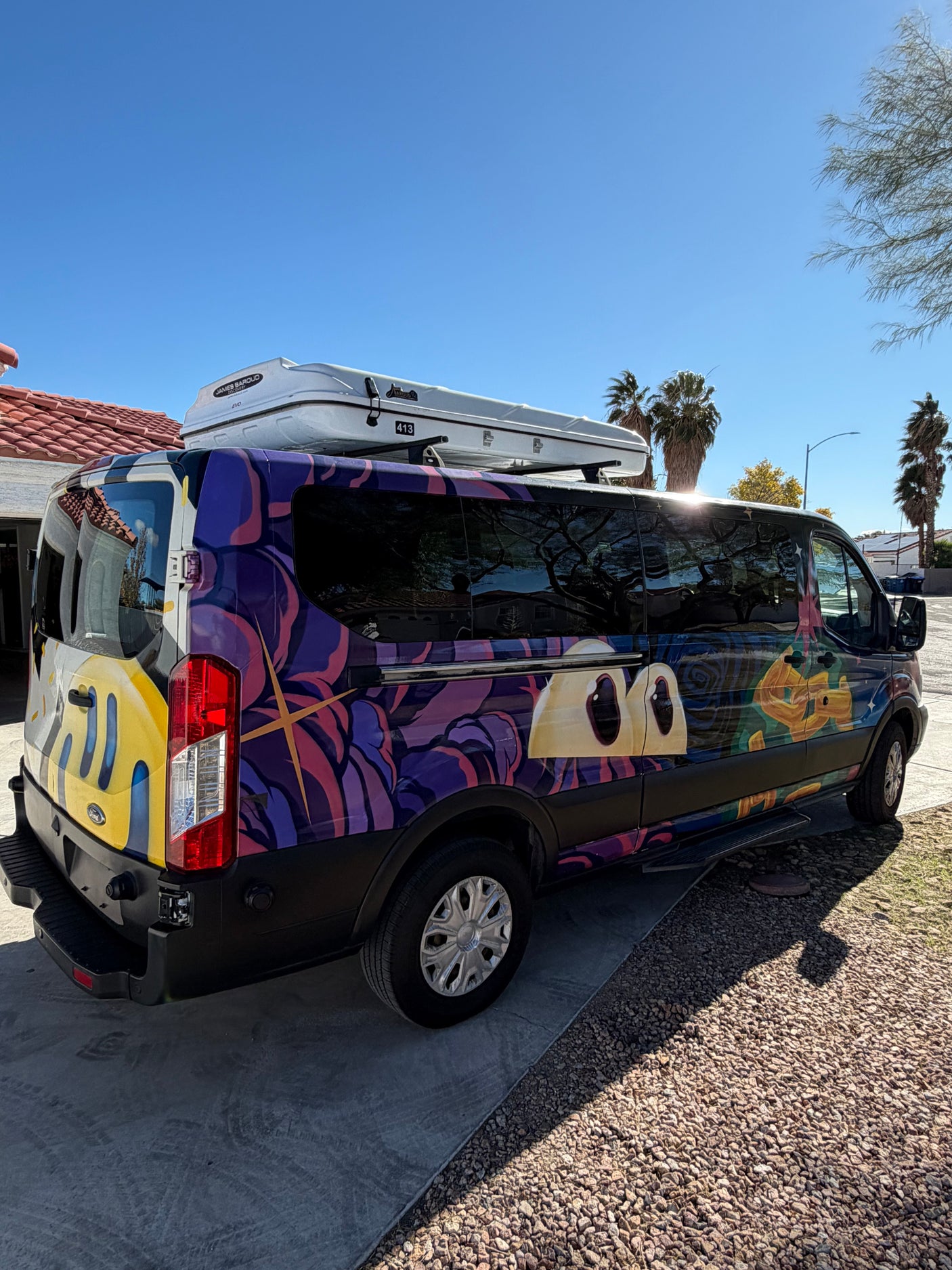 A colorful custom-wrapped RV rental van with abstract art, large eyes, and a rooftop cargo box, parked on a sunny day.