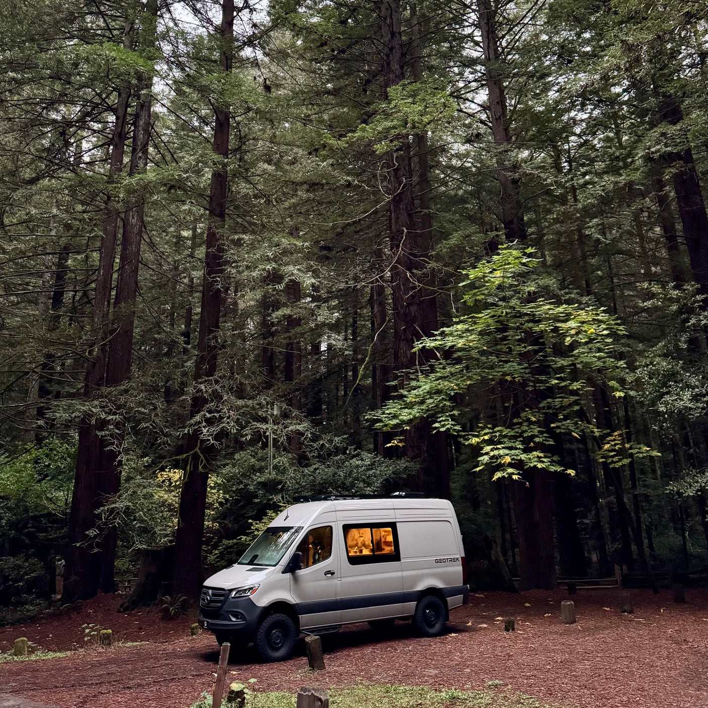 An RV rental, a white GEOTREK camper van, is nestled in a dense redwood forest at dusk, its interior warmly lit.