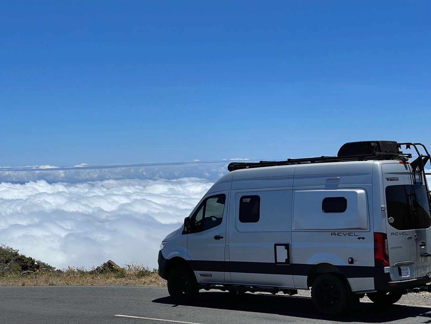 White Winnebago Revel 4x4 camper van on a mountain road, high above clouds. Blue sky. Epic rv rental adventure awaits!