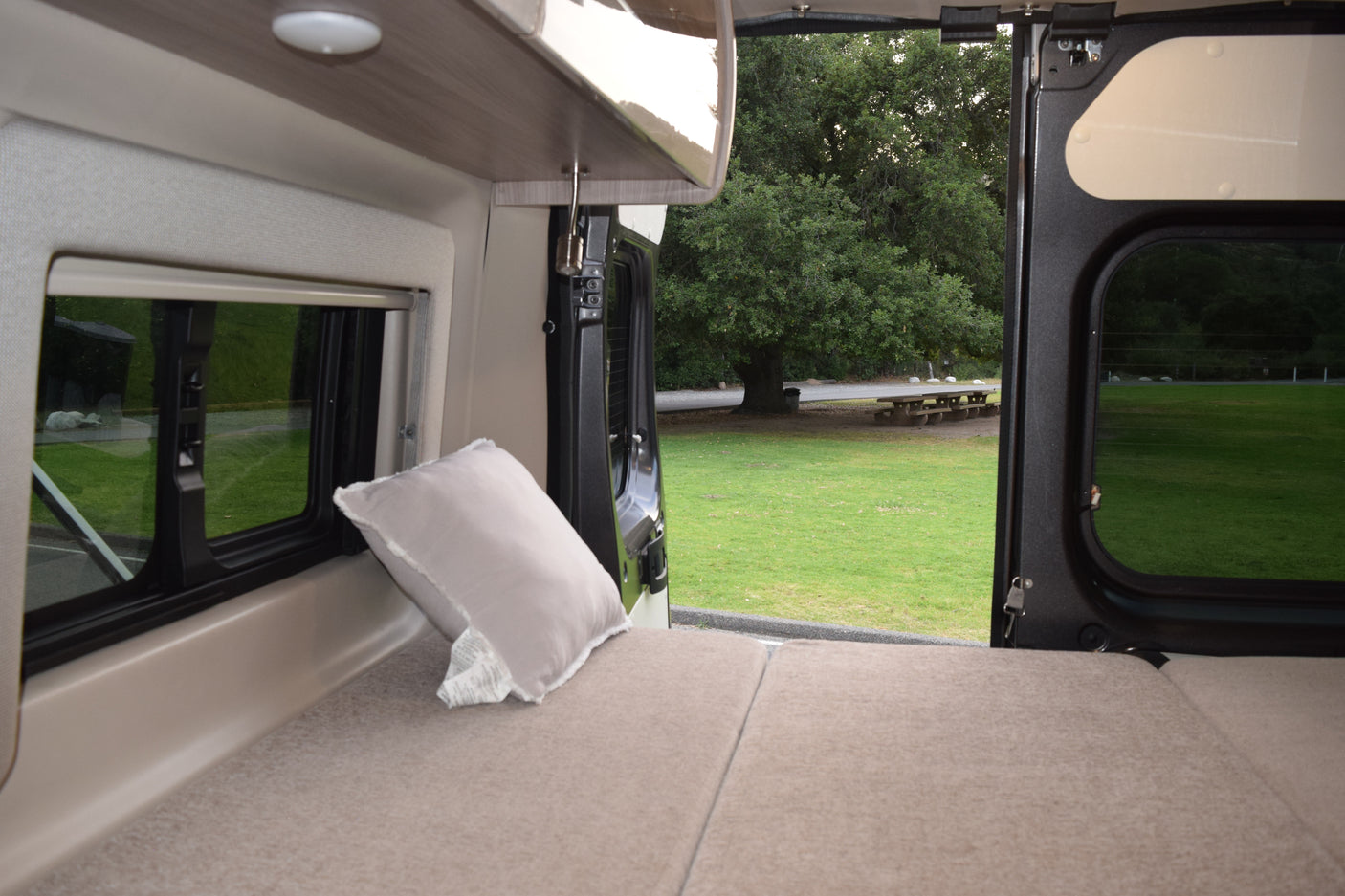 Experience RV rental comfort: cozy bed inside, scenic park view with a large tree & picnic table right outside.