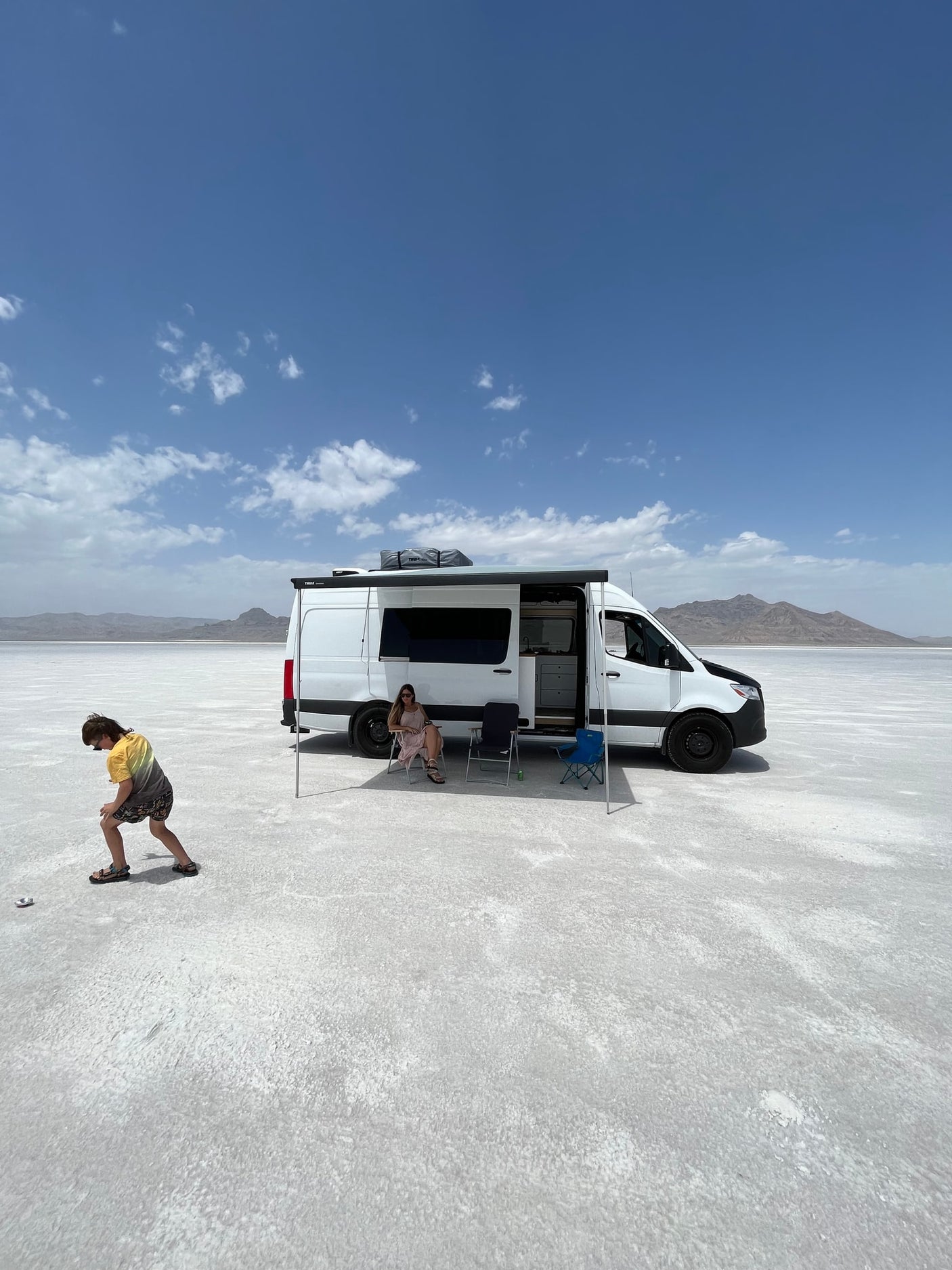 RV rental on vast salt flats under a blue sky. A woman relaxes under the awning, a child plays nearby. Road trip fun!