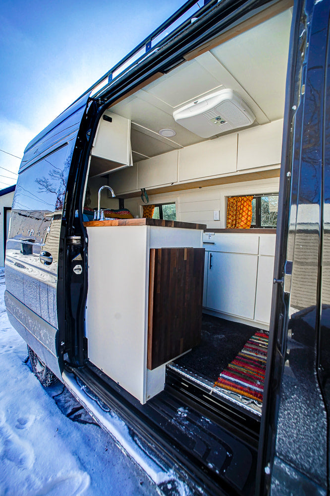 This rv rental camper van's custom kitchen and living area, with a sink, wooden counters, and cabinets, seen from its open side door in the snow.