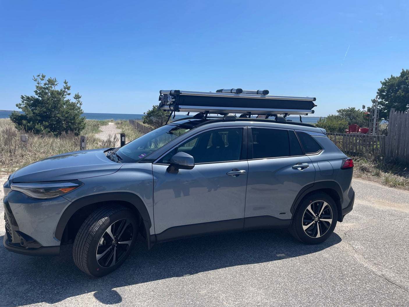 A grey Toyota Corolla Cross with a rooftop tent is parked by a sunny beach. Ideal for outdoor adventures, like an RV rental alternative.
