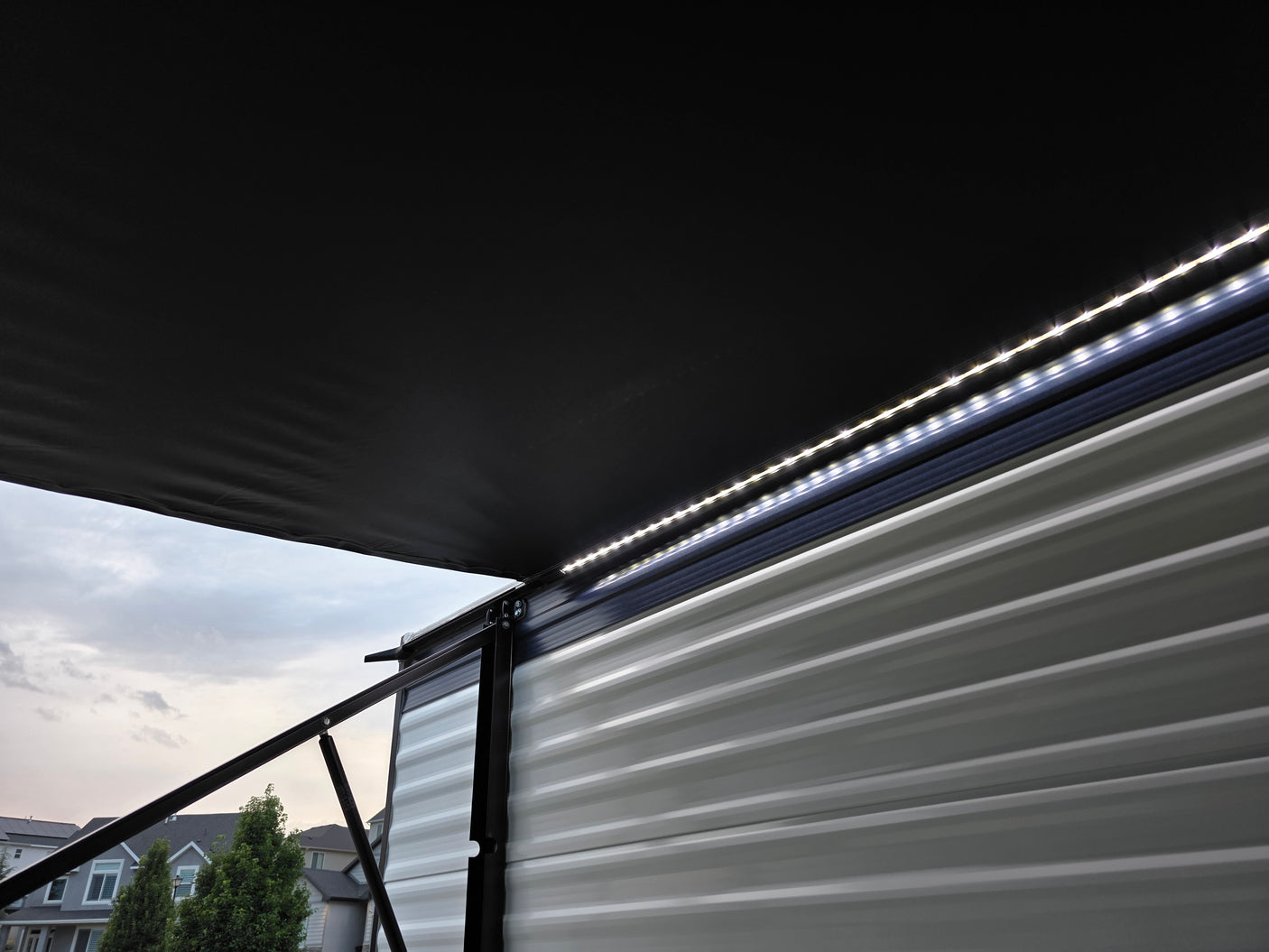 This close-up of an RV rental shows a black extended awning with bright LED strip lights illuminating the ribbed side paneling.