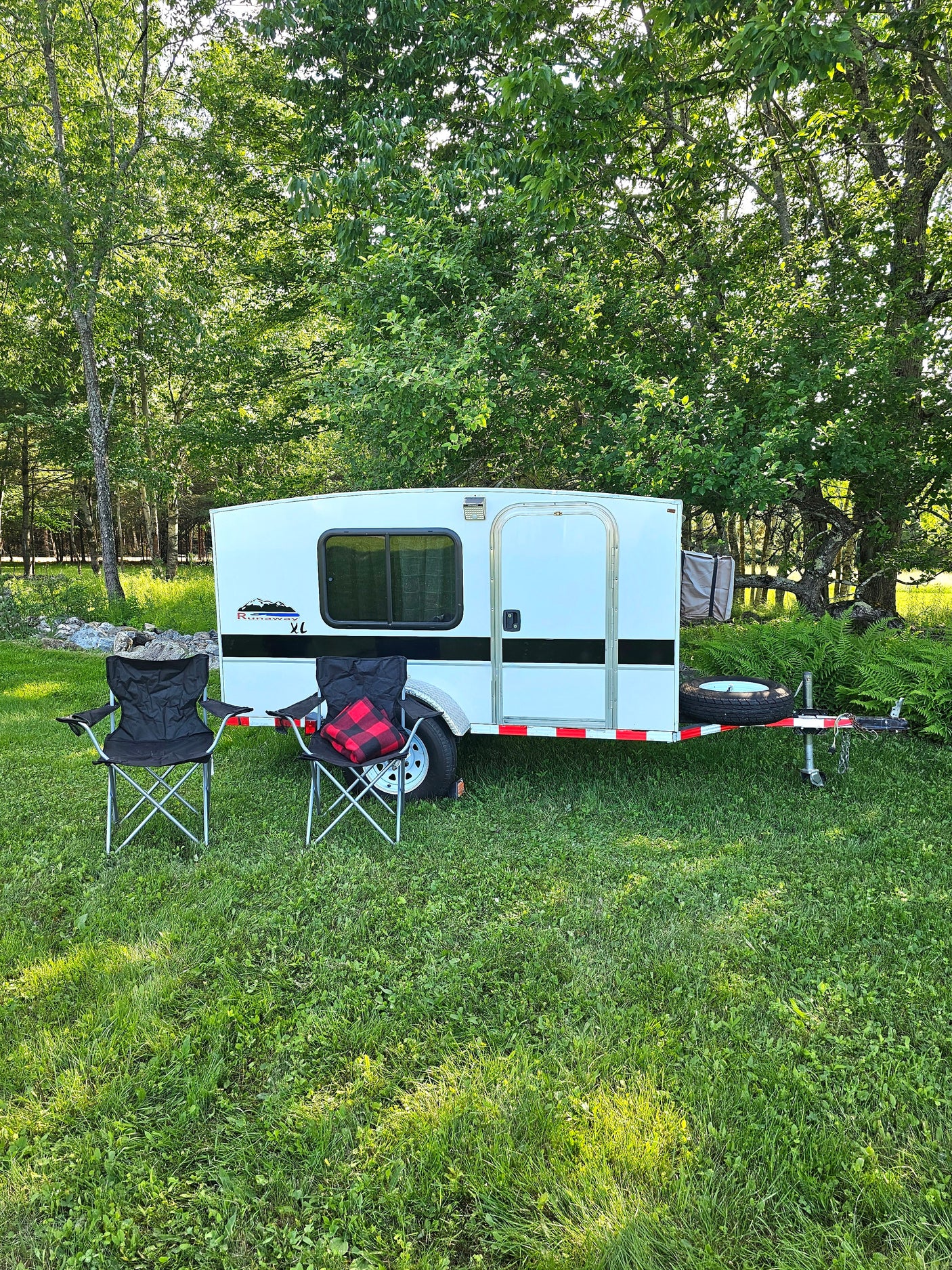 A white camper trailer, like an rv rental, with two folding chairs in a sunny, grassy spot under trees. Ready for your getaway!