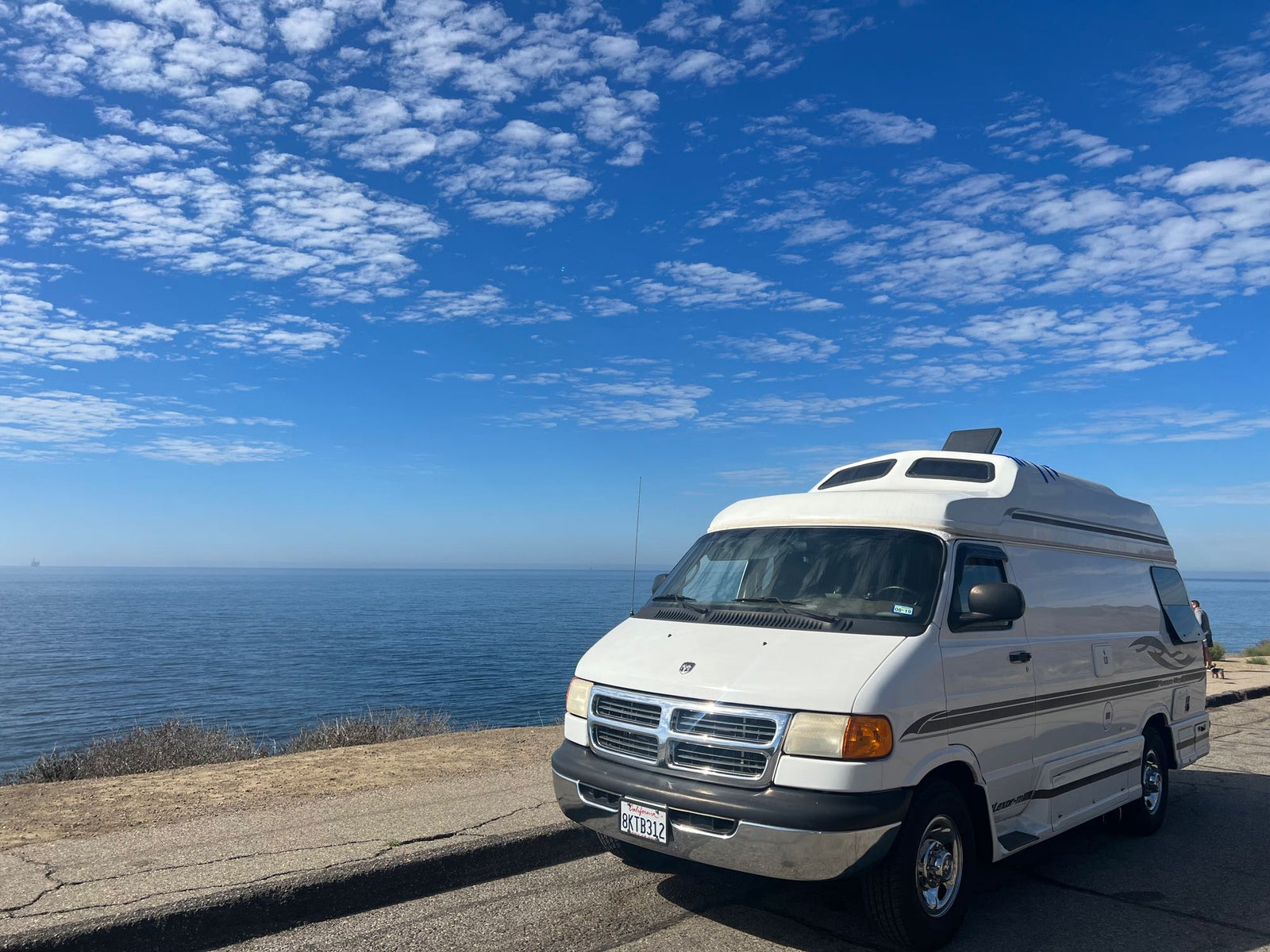 A white camper van, perfect for an RV rental adventure, parked by the ocean under a blue sky with fluffy clouds.
