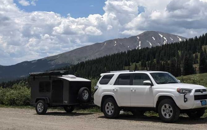 A white SUV with an off-road RV rental camper is perfect for mountain adventures. Scenic backdrop with forests & snowy peaks.
