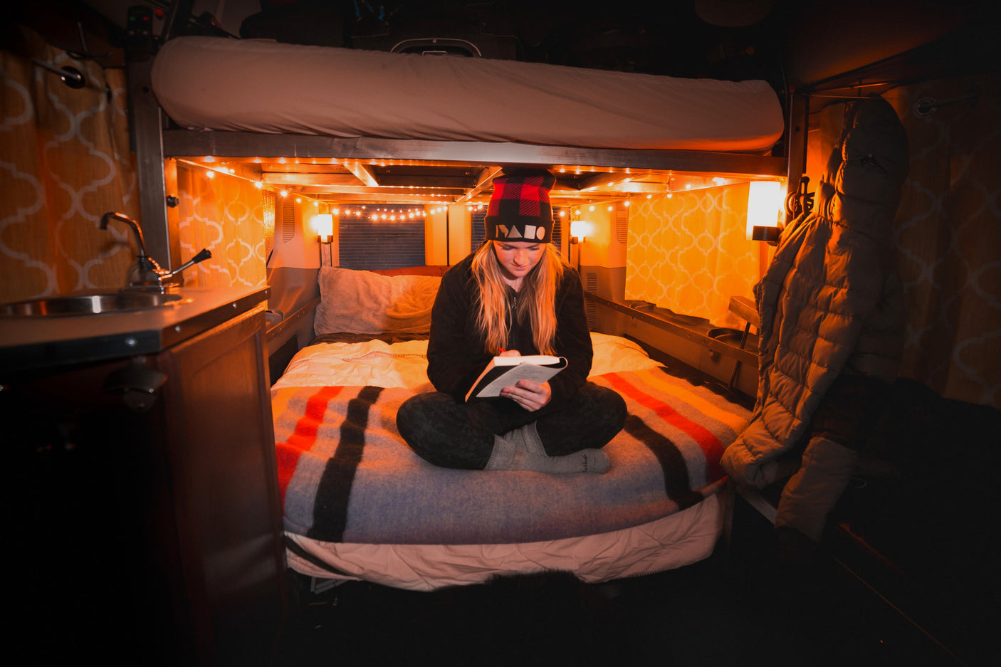 Cozy RV rental interior: A woman reads on a comfortable bed under warm string lights, featuring a bunk bed above and small sink area.