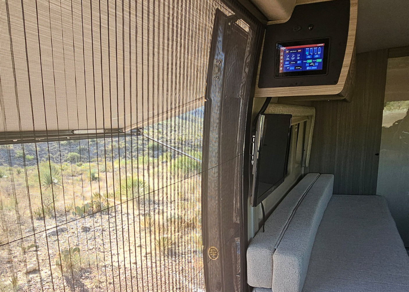 Inside this RV rental: a large window with desert landscape, overhead control panel, TV, and bench seating.