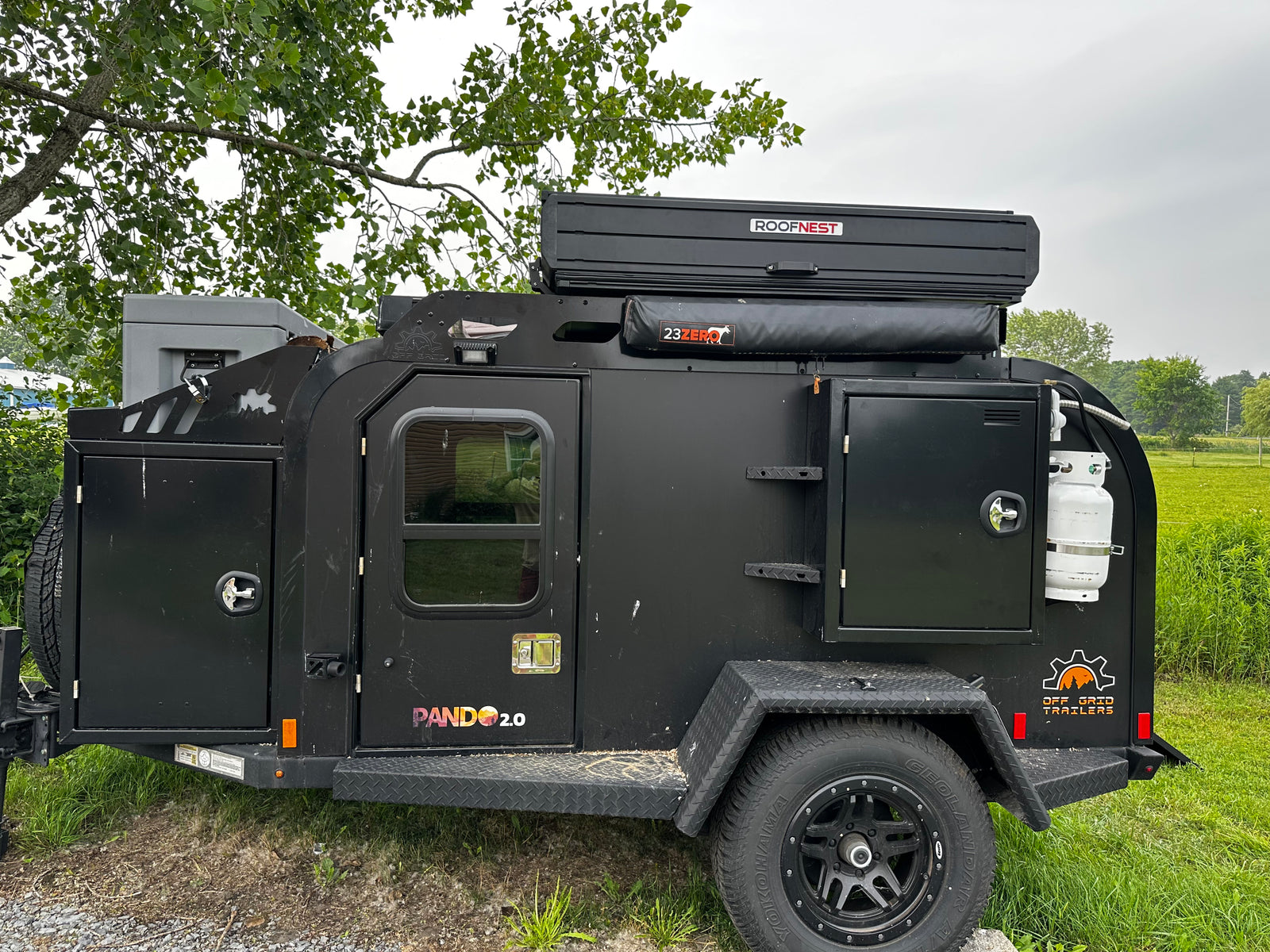 parked at an RV rental park under tall trees.|Black Jeep Gladiator towing an Off Grid overland trailer with a Roofnest tent