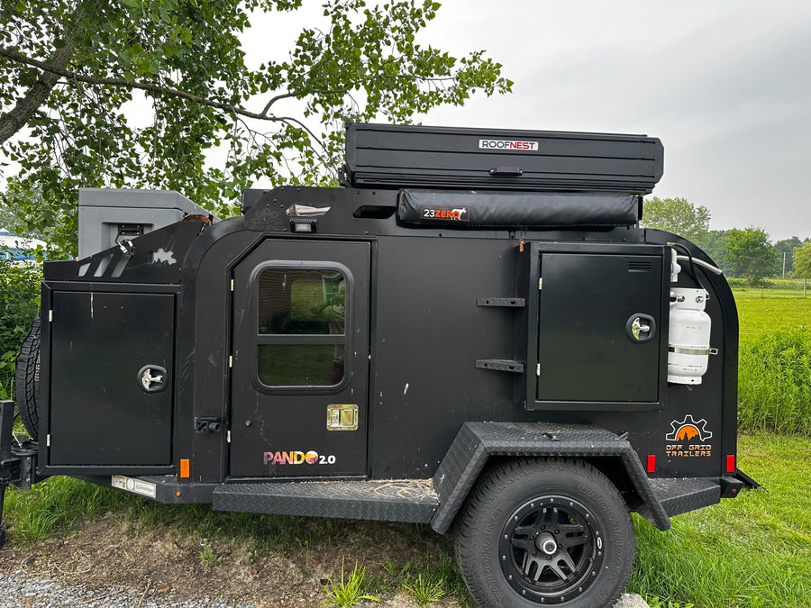 parked at an RV rental park under tall trees.|Black Jeep Gladiator towing an Off Grid overland trailer with a Roofnest tent