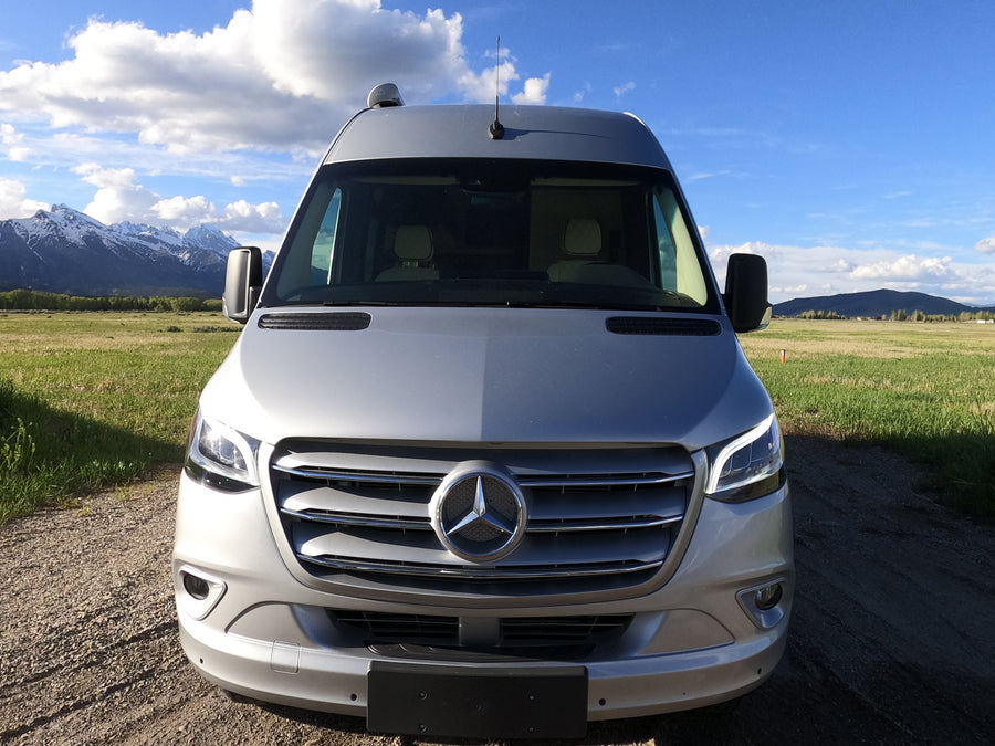 Silver Mercedes-Benz Sprinter camper van RV rental on a gravel road, backed by majestic snow-capped mountains and green fields.