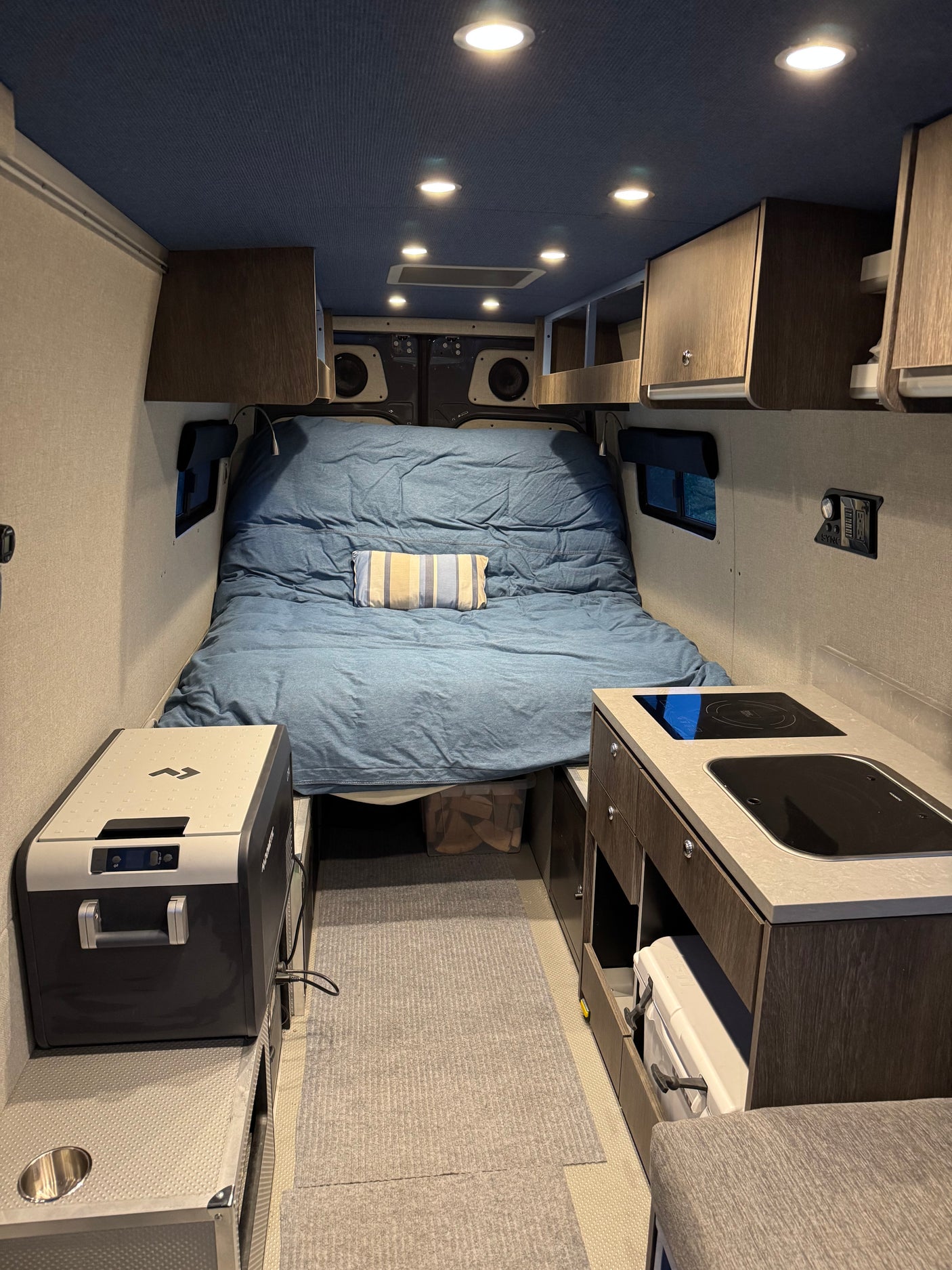 This rv rental interior features a comfortable bed, compact kitchen with cooktop, and a portable fridge, all within a well-lit space.