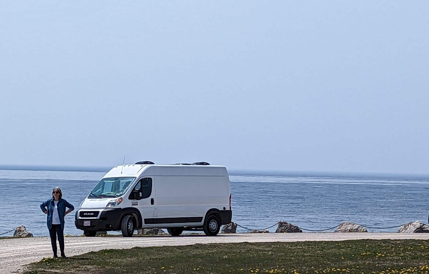 A white RAM ProMaster van, likely an RV rental, is parked near a calm blue body of water. A person stands on the left.