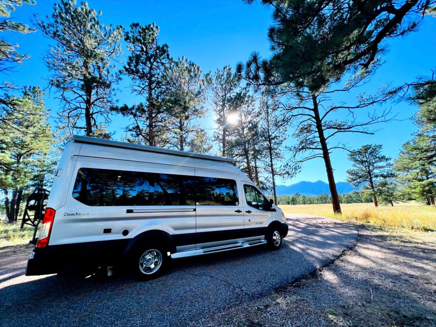 This silver rv rental camper van is parked on a road surrounded by sunny pine trees & distant mountains, ready for adventure!