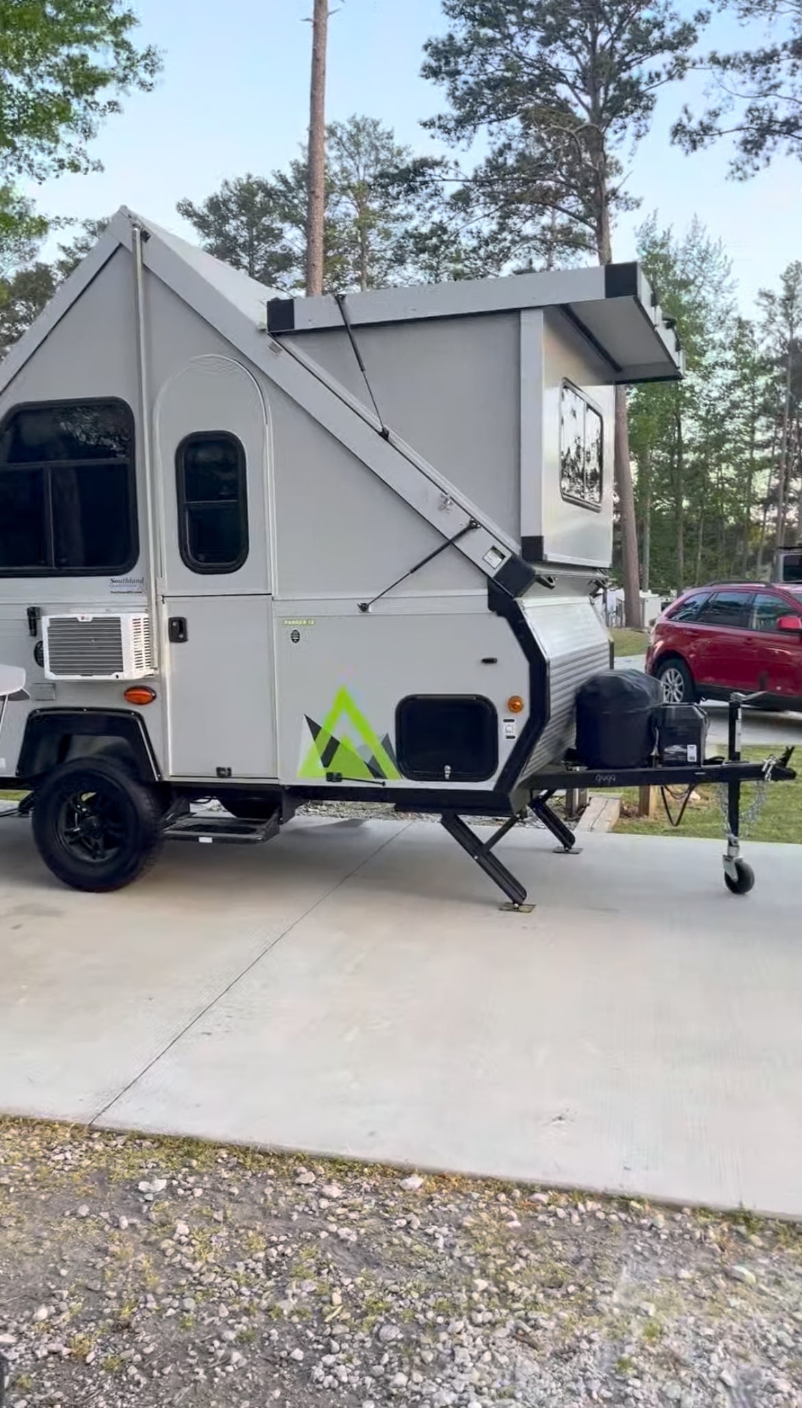 A light gray A-frame pop-up camper, a great rv rental, set up on concrete in a wooded campsite.