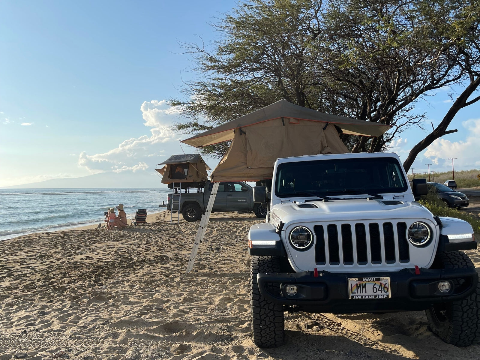 White Jeep with rooftop tent (RV rental option) parked on a sandy beach. People relax by the ocean. Ideal for beach camping adventures.