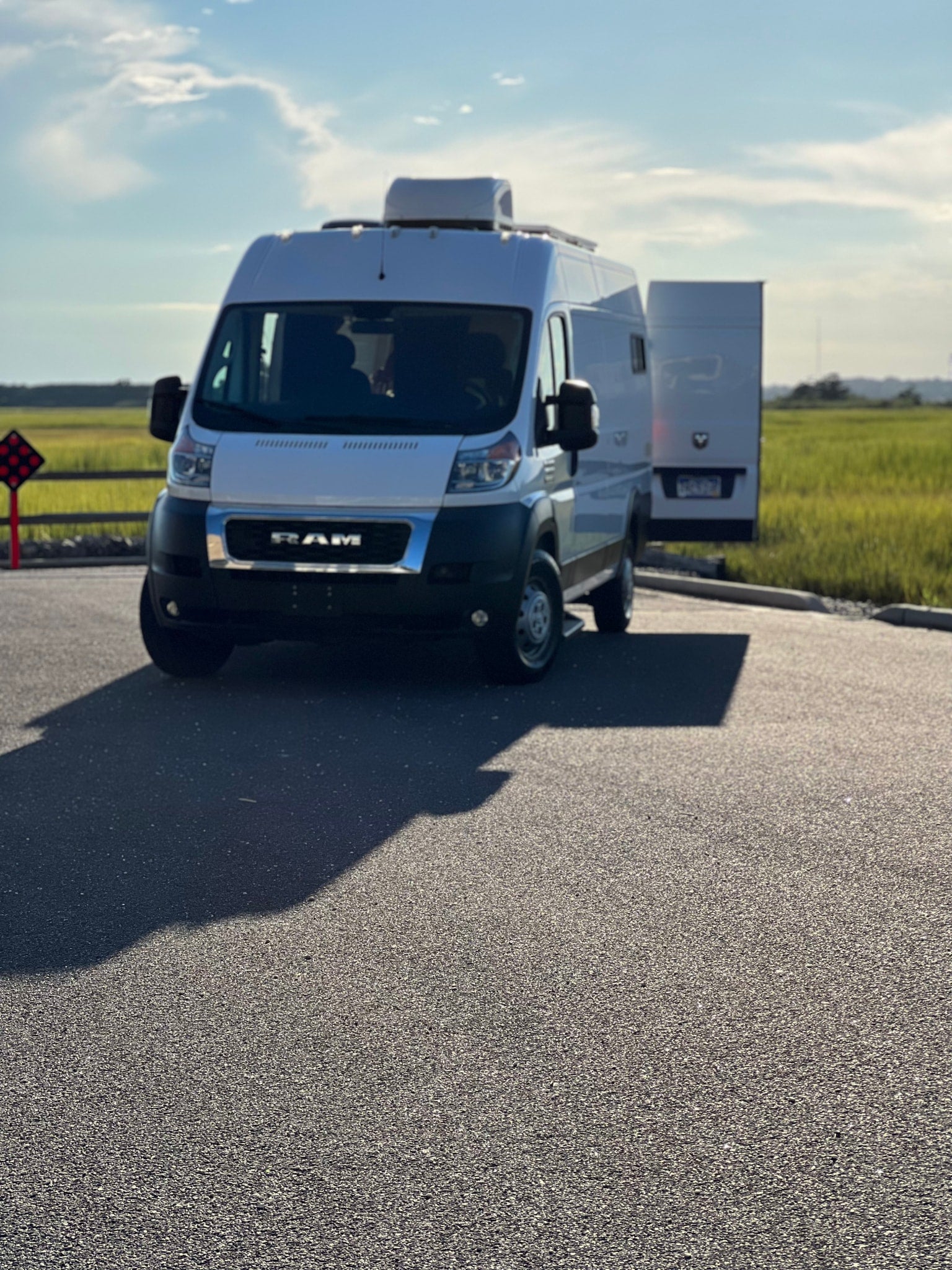 White Ram ProMaster RV rental van parked in a sunny lot, ready for adventure. Grassy field and blue sky in background.