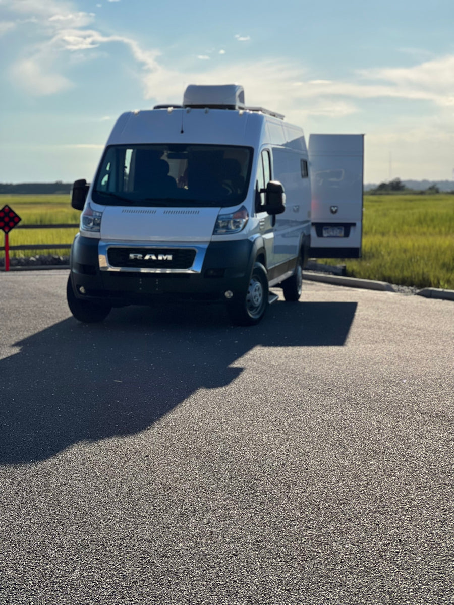 White Ram ProMaster RV rental van parked in a sunny lot, ready for adventure. Grassy field and blue sky in background.