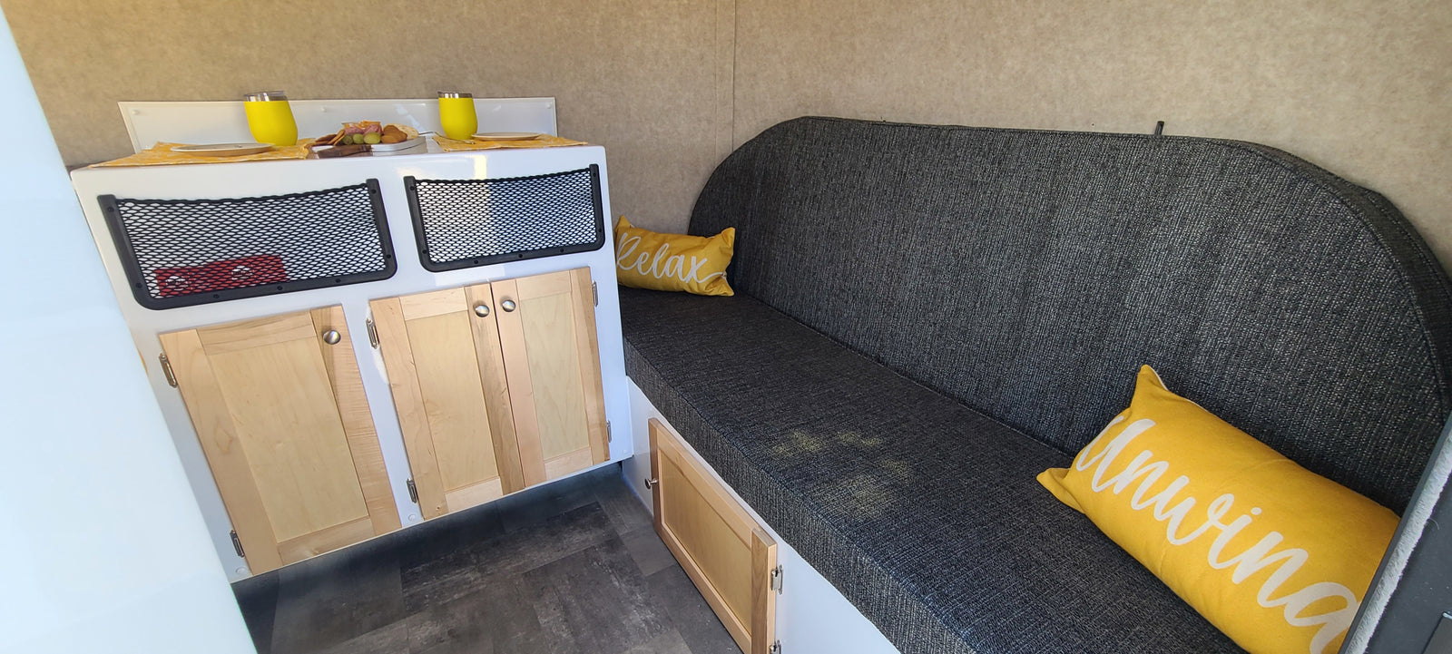 This rv rental features a cozy grey sofa with yellow pillows, a compact kitchenette with snacks, and storage cabinets.