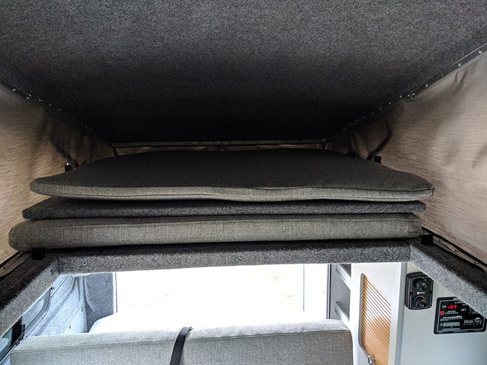 RV rental pop-top bed shows stacked cushions for comfortable sleeping. Elevated interior view of a camper.