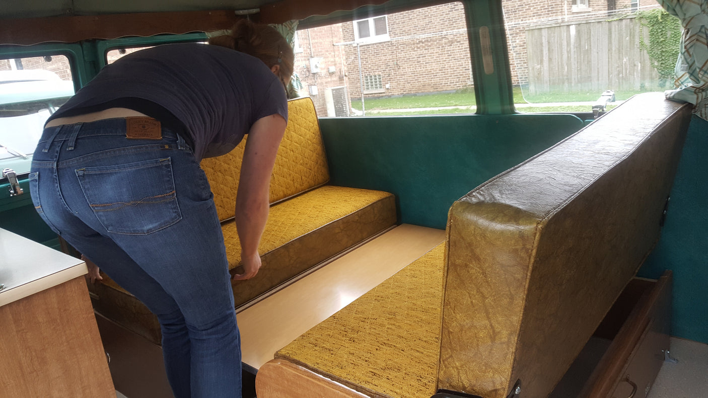 Inside an **RV rental**, a person in blue jeans is seen bending, converting the yellow benches into a sleeping area.