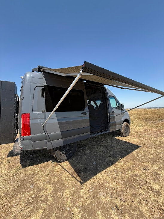 Grey off-road camper van with an extended awning in a dry field, ready for adventure. Perfect for RV rental.