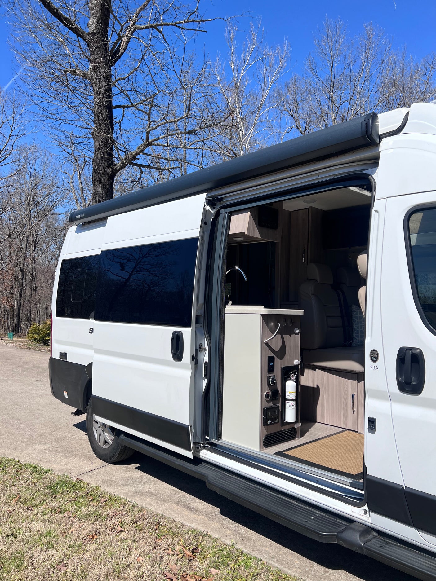 White RV rental camper van with open side door, revealing interior kitchen, seating, and a fire extinguisher. Awning extended.