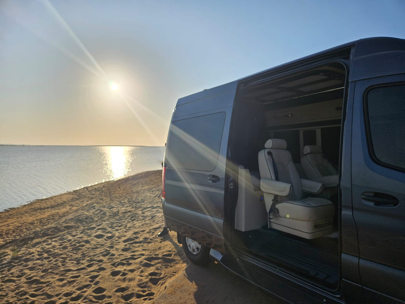 Luxurious RV rental parked on a sandy beach at sunset, showcasing plush interior seats by the tranquil water.